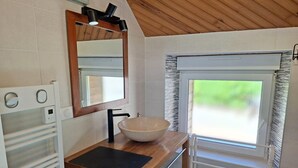 Ein Badezimmer mit einem Holzbadezimmer, einem großen Spiegel, einem Waschbecken, einem Handtuchhalter und einem Fenster mit Blick auf Grünflächen.