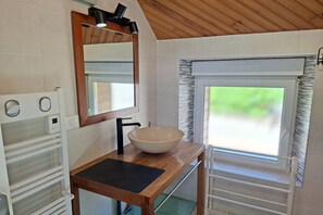A bathroom with a wooden vanity, a large mirror, a vessel sink, a towel rack, and a window with a view of greenery. - Breton Stone Cottage by Sandy Beaches (Landéda)
