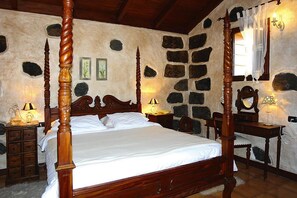 A four-poster bed with a wooden headboard, a stone wall with embedded rocks, a wooden nightstand with a lamp, and a wooden dresser with a mirror. - Seaview Holiday Home for 4 (Vera de Erques)