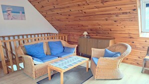 Ein gemütliches Zimmer mit Holzbalkendecke, einem Korbsessel mit blauen Kissen, einem passenden Sessel, einem kleinen Holztisch und einem Schrank.
