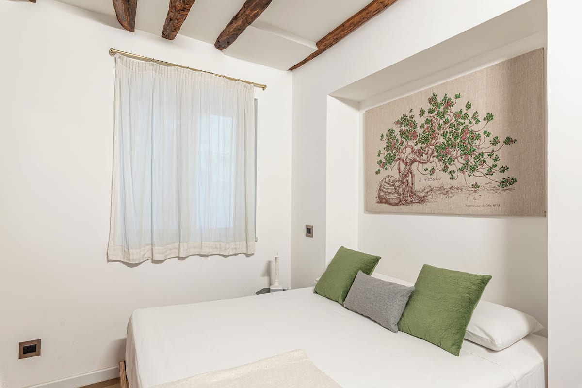Um quarto com cama, janela com cortinas, quadro na parede e mesa de cabeceira com abajur.