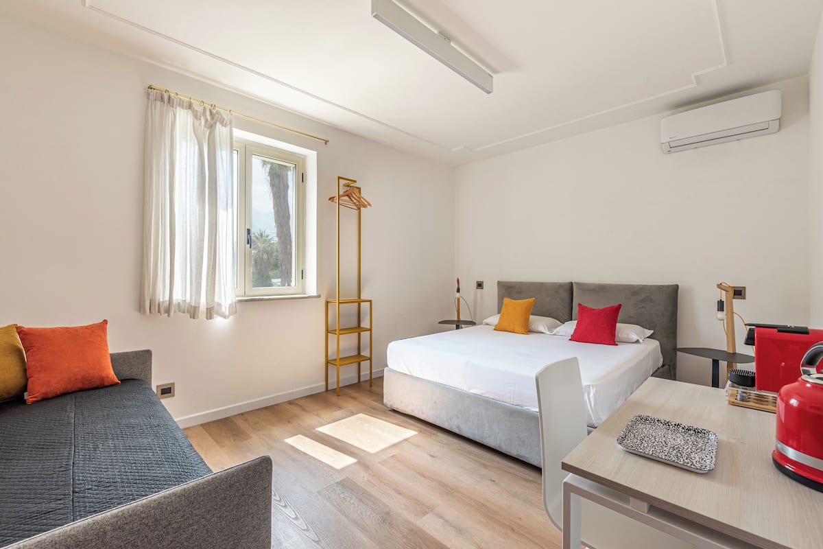 Quarto de hotel com uma cama, escrivaninha, cadeira, guarda-roupa e janela.
