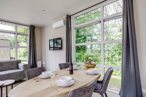 A dining area with a wooden table set for four, large windows, a grey sofa, and a flat-screen TV mounted on the wall. - Lodge 18 | 4 persoons chalet in Zennewijnen (Zennewijnen)