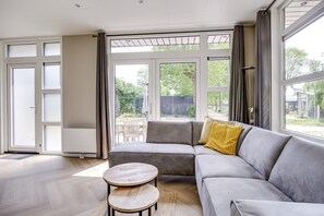 Ein modernes Wohnzimmer mit einer grauen Eckcouch, einem runden Couchtisch und Blick auf einen Garten durch große Fenster.