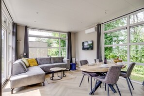 A modern living room with a grey sectional sofa, a round dining table with chairs, and large windows offering a view of greenery. - Lodge 18 | 4 persoons chalet in Zennewijnen (Zennewijnen)