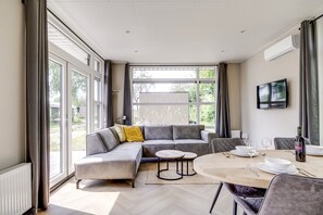 A modern living room with a grey sectional sofa, a round dining table, and a TV mounted on the wall. - Lodge 18 | 4 persoons chalet in Zennewijnen (Zennewijnen)