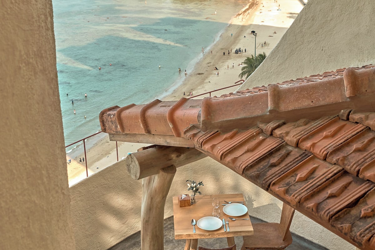 A balcony with a terracotta roof, a table with a vase, and a view of a beach and ocean.