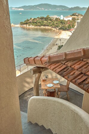 Uma varanda com telhado de terracota, uma mesa com um vaso e vista para a praia e o oceano.