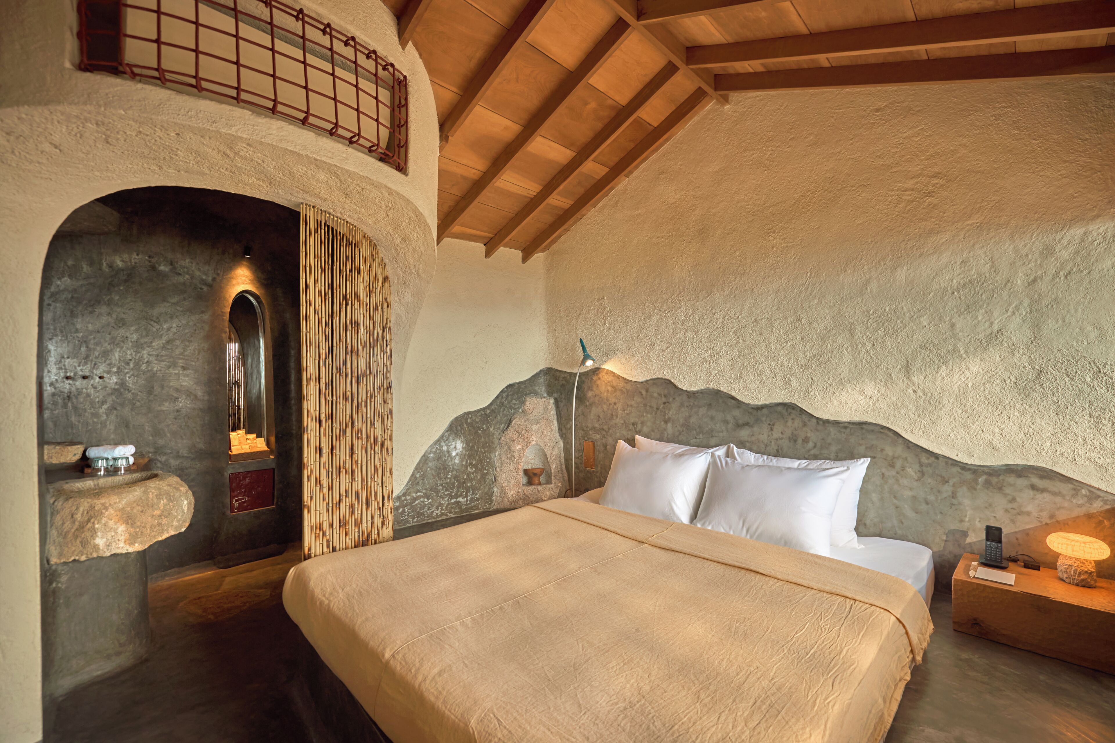 A bedroom with a stone wall, a bed with white linens, a nightstand with a lamp, and a small window with a view.