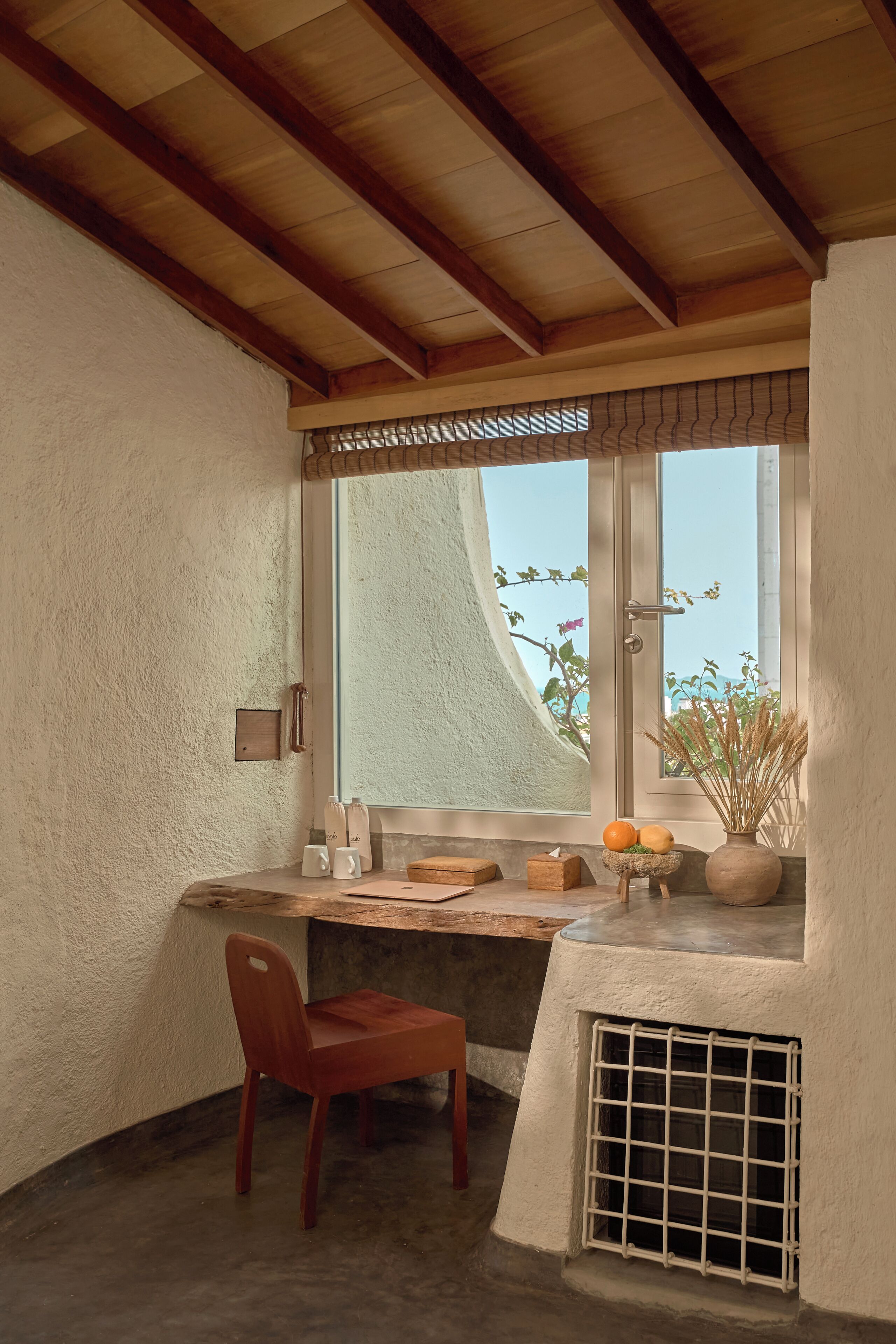 A corner room with a wooden ceiling, a window with a view of plants, a chair, a small table with a vase and oranges, and a vent.
