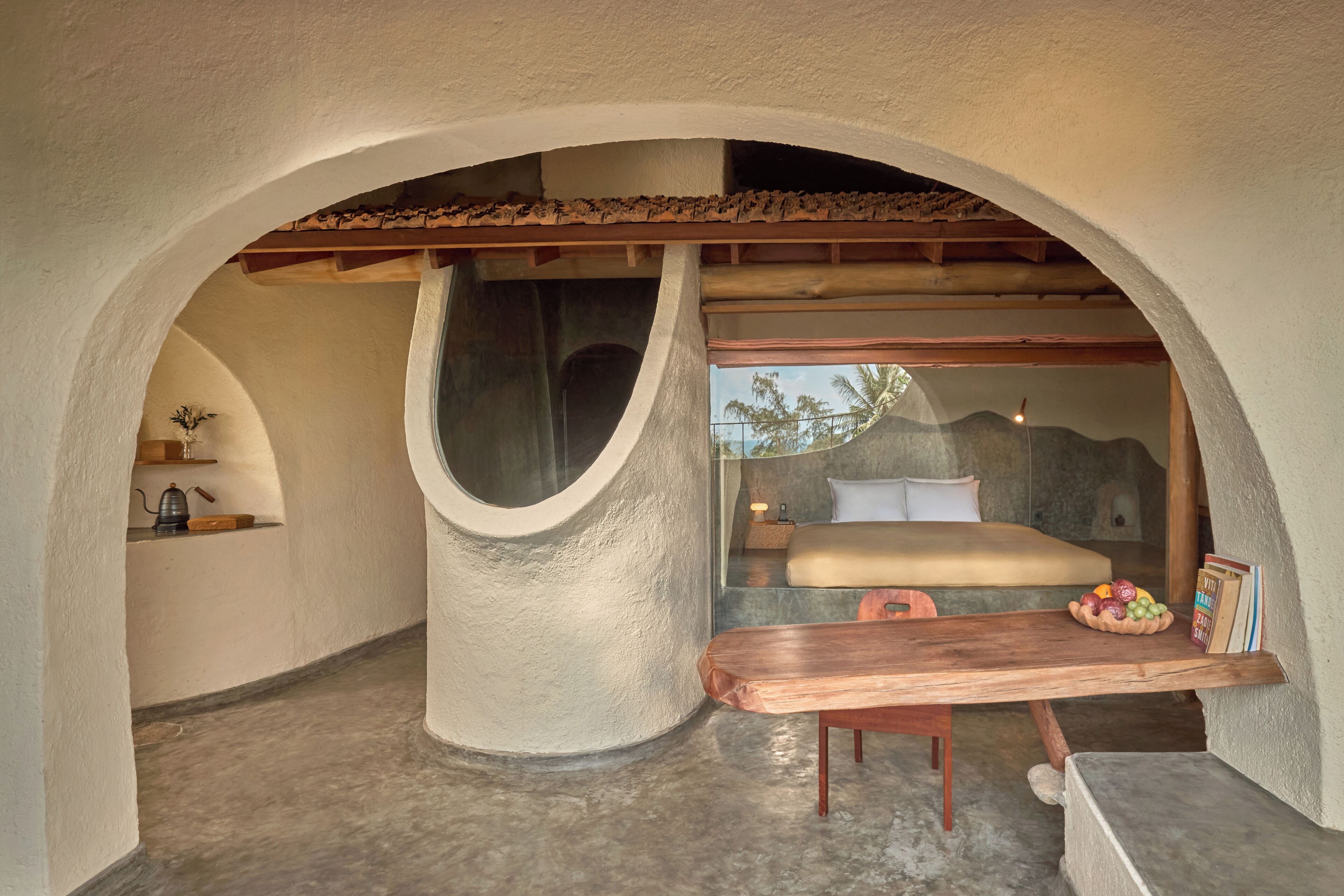 A cozy bedroom with a bed, a wooden table, and a window overlooking a tropical landscape.