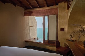 Quarto com cama, mesa de madeira, cadeira, janela com vista para o mar e uma planta.
