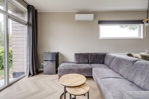 Una sala moderna con un sofá seccional gris, una pequeña mesa de madera, un equipo de aire acondicionado integrado y un ventanal con cortinas.