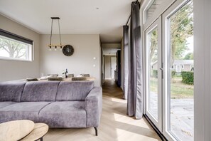A modern living room with a grey sofa, a dining table with chairs, a hanging light fixture, and a view of the outdoors through large windows.