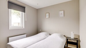 A bedroom with two single beds, a window with a blind, a bedside table with a lamp, and two framed pictures on the wall.