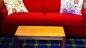 Un canapé rouge agrémenté de coussins à motifs floraux, une table basse en bois et une étagère ornée d’assiettes décoratives et d’une statuette.
