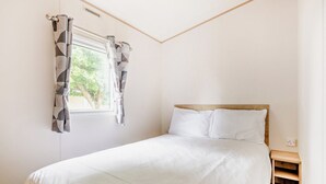 A bedroom with a bed, a window with curtains, a wooden headboard, and a nightstand. - Gold 3 Bedroom Caravan, Sleeps 8. With parking. (Great Yarmouth)