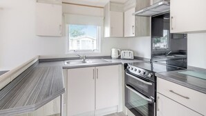 A modern kitchen with white cabinetry, a stainless steel range hood, a double oven, and a window with a view of a house. - Gold 3 Bedroom Caravan, Sleeps 8. With parking. (Great Yarmouth)
