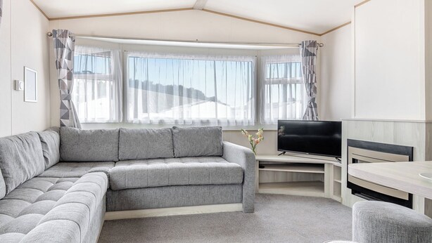 A modern living room with a grey sofa, a flat-screen TV, and a dining area. - Gold 3 Bedroom Caravan, Sleeps 8. With parking. (Great Yarmouth)