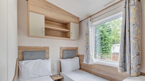Una habitación con dos camas, un pequeño estante de madera, una ventana con cortinas y un espejo sobre el estante.