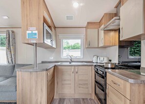 A compact kitchen with wooden cabinets, a built-in microwave, a stove, and a window providing natural light. - Gold Caravan. With parking and decking. (Kidwelly, Carmarthenshire)