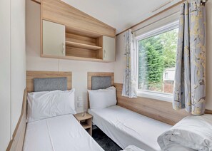 A room with two beds, a small wooden shelf, a window with curtains, and a mirror above the shelf. - Gold Caravan. With parking and decking. (Kidwelly, Carmarthenshire)