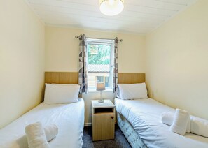 A room with two beds, a small bedside table, a window with curtains, and a ceiling light. - Superior Lodge. With parking, ensuite and decking. (Great Yarmouth)