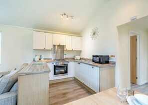 A compact kitchen with white cabinets, a built-in oven, a microwave, and a dining area with a wooden table and chairs. - Superior Lodge. With parking, ensuite and decking. (Great Yarmouth)