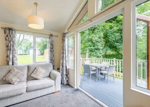 A room with a sofa, a hanging light fixture, a sliding glass door leading to an outdoor deck with a table and chairs. - Superior Lodge. With parking, ensuite and decking. (Great Yarmouth)