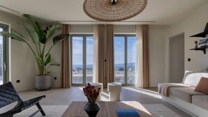 Un salon moderne avec une grande plante en pot, un fauteuil noir en osier, une table basse en bois et un canapé offrant une vue dégagée grâce à de grandes fenêtres.