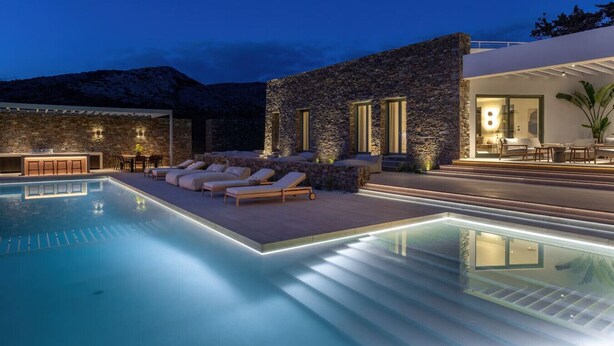 Un espace aménagé au bord de la piscine, avec des chaises longues, un bar et un bâtiment en pierre doté de grandes fenêtres.