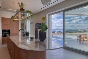 A modern kitchen with a curved island, large windows, and a view of a landscape. - Villa Calisto – A Sunset Sanctuary (2524+VXR Paros, Greece)