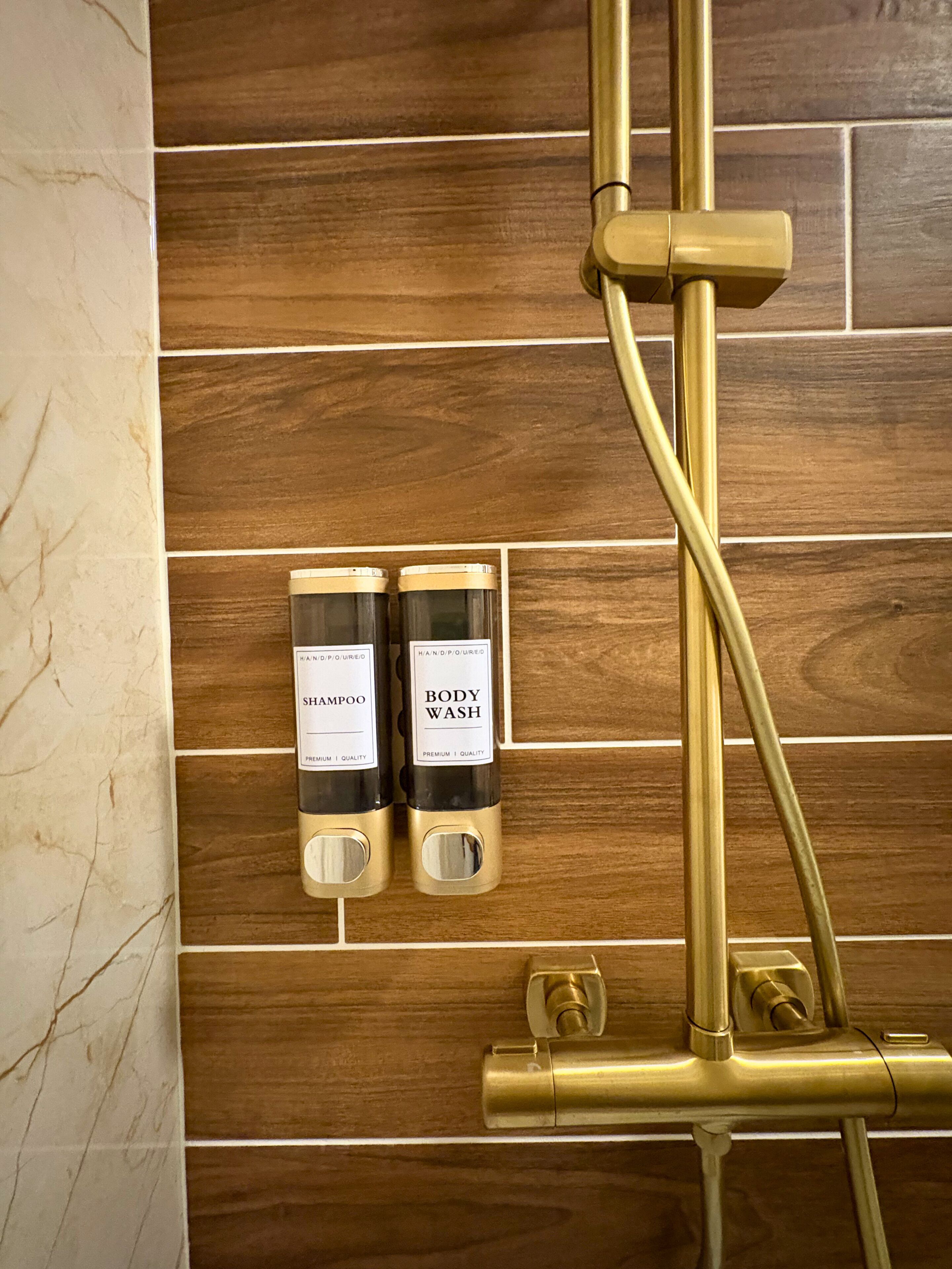A bathroom with a wooden wall, two bottles of shampoo and body wash, and a gold-colored shower system.