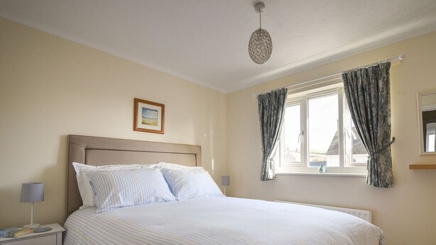 A bedroom with a bed, a nightstand, a lamp, a window with curtains, and a picture on the wall. - Mead Cottage (Bridport)