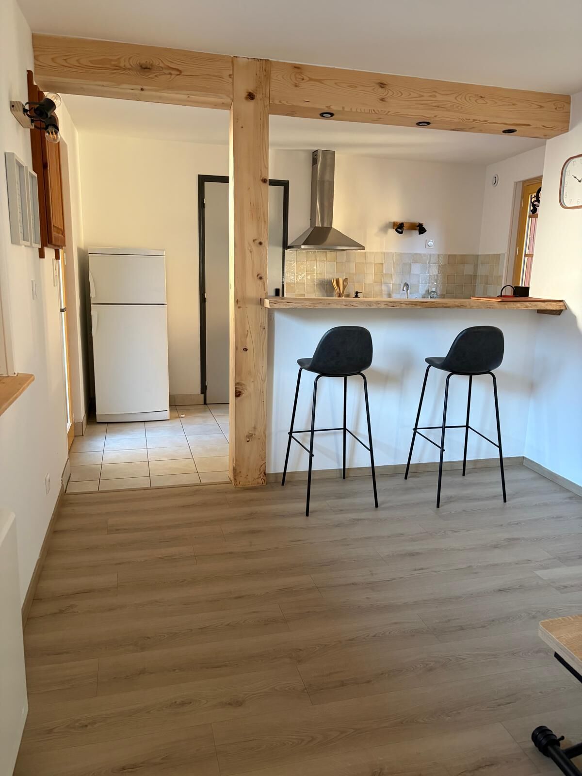 Une cuisine avec un plafond orné de poutres en bois, deux tabourets de bar noirs, un réfrigérateur blanc et un mur recouvert de carreaux.