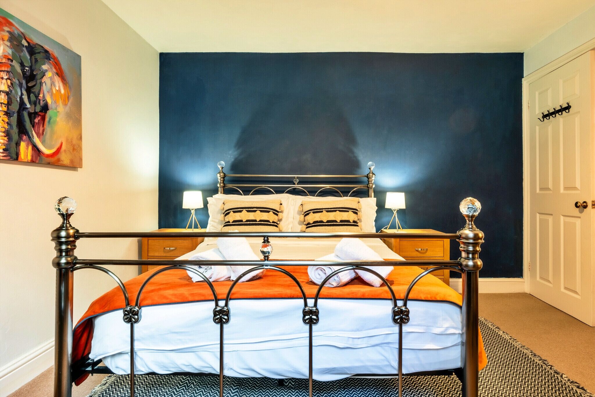 A bed with a metal frame, two pillows, a white and orange bedspread, and a dark blue wall behind it.