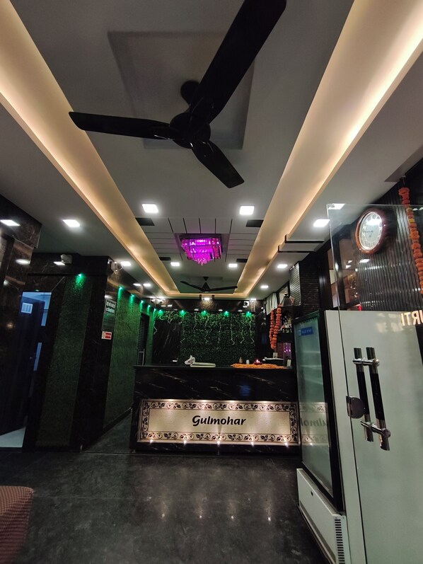 Eine Hotellobby mit Deckenventilator, eingelassener Beleuchtung, einem Ziertelegramm, einer Rezeption und einem Kühlschrank.