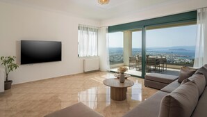 A spacious living room with a large flat-screen TV, a comfortable sofa, a round coffee table, and a view of a cityscape through a large window. - Lithea Estate Collection (Chania)