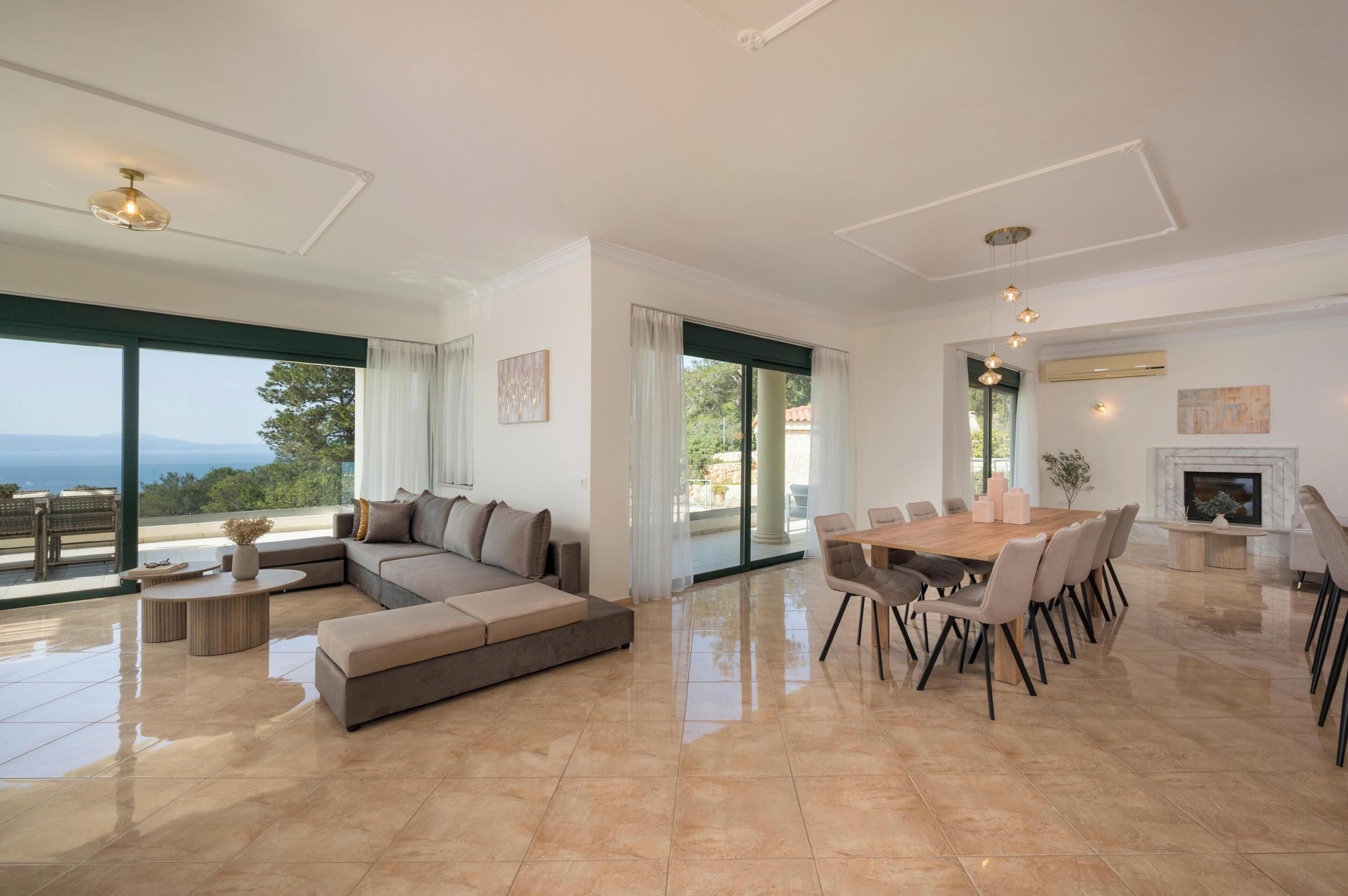 A spacious living area with a large sofa, a dining table with chairs, a fireplace, and large windows offering a view of the outdoors.