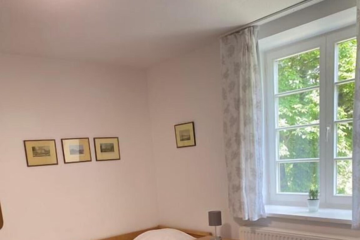 Una cama individual con estructura de madera, una mesita de noche pequeña, un radiador y una ventana con cortinas.