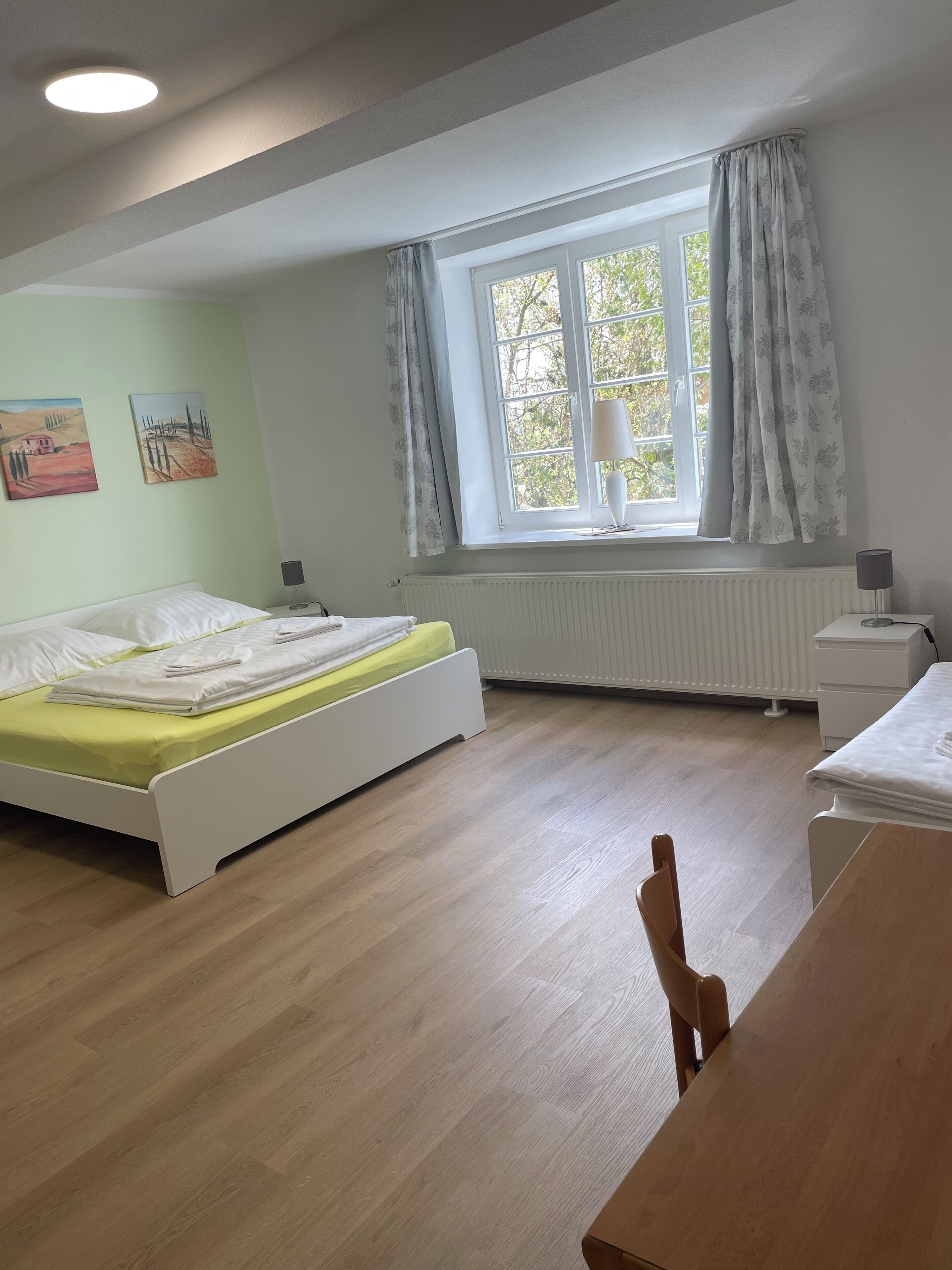 A room with a bed, a table, a chair, a window with curtains, a radiator, and wall art.