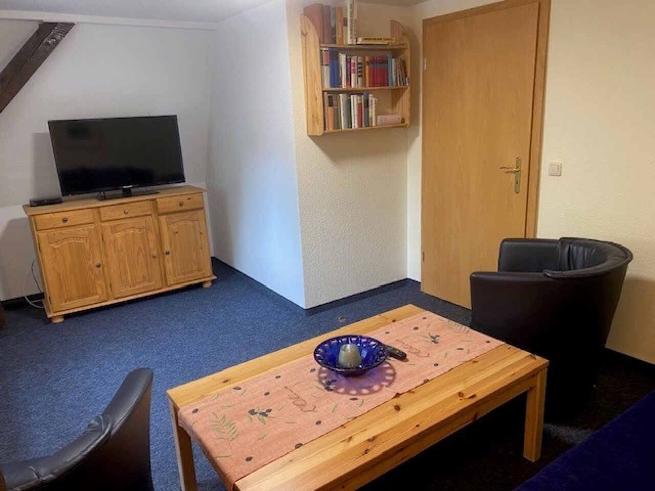 Une pièce comprenant un téléviseur posé sur un meuble en bois, une bibliothèque, une table avec un bol et une poubelle.