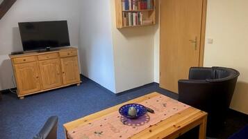 Une pièce comprenant un téléviseur posé sur un meuble en bois, une bibliothèque, une table avec un bol et une poubelle.