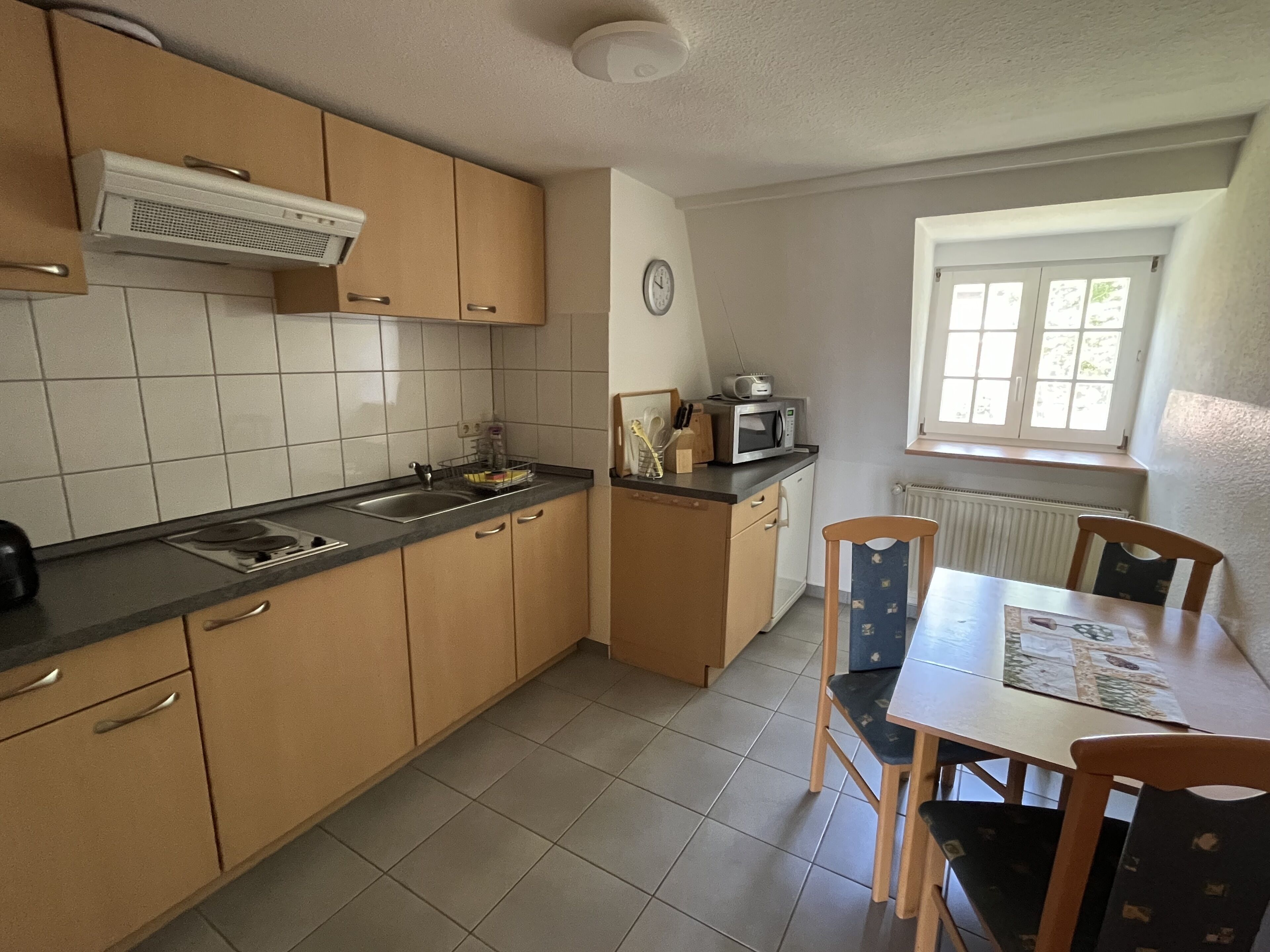 A compact kitchen with a stove, sink, and microwave. There is a dining table with chairs and a window with blinds.
