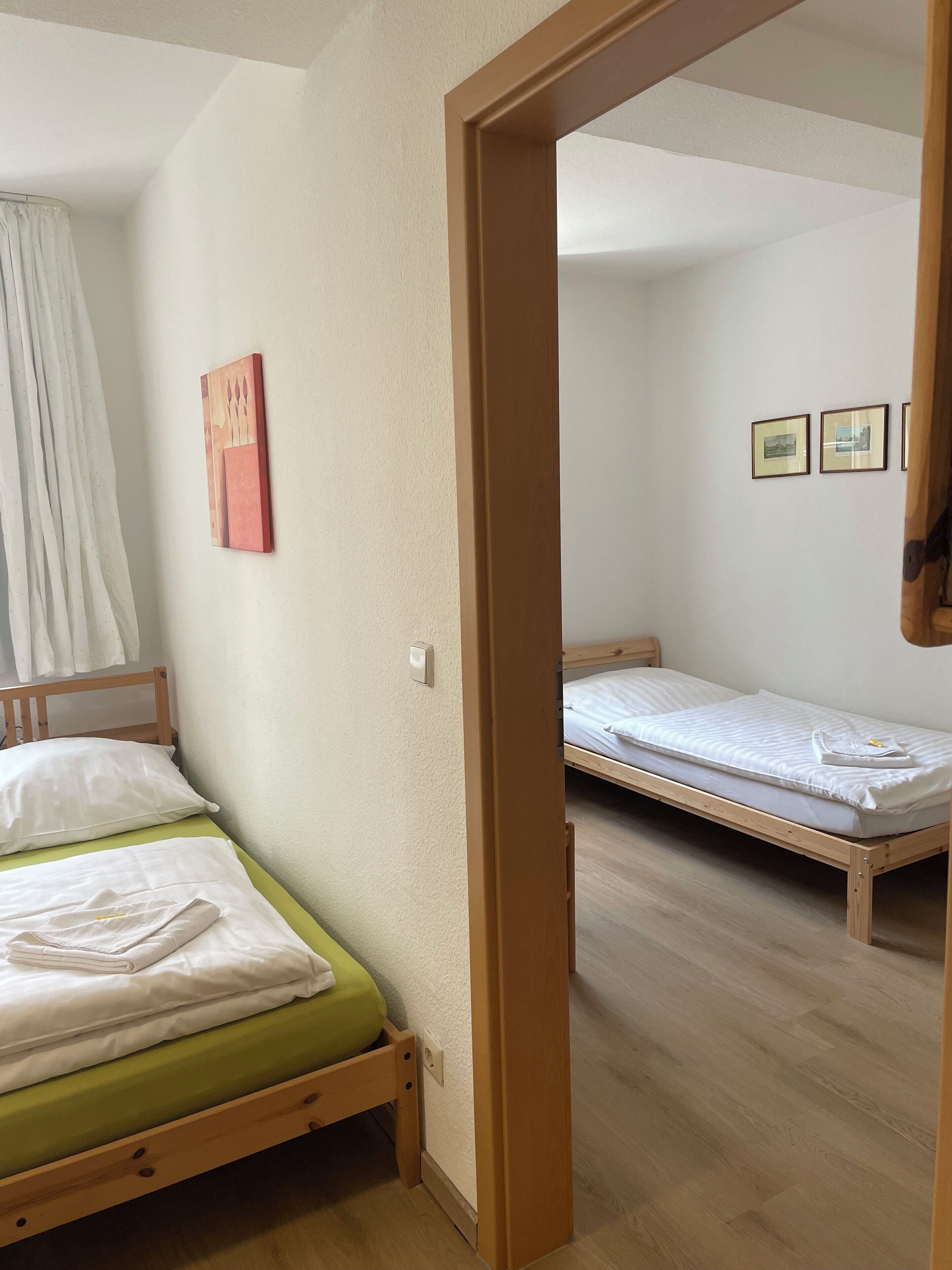 A room with two wooden beds, a green mattress on one bed, and a white mattress on the other. There is a white curtain partially covering a window, and a framed picture on the wall.