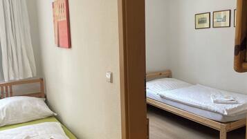 A room with two wooden beds, a green mattress on one bed, and a white mattress on the other. There is a white curtain partially covering a window, and a framed picture on the wall.