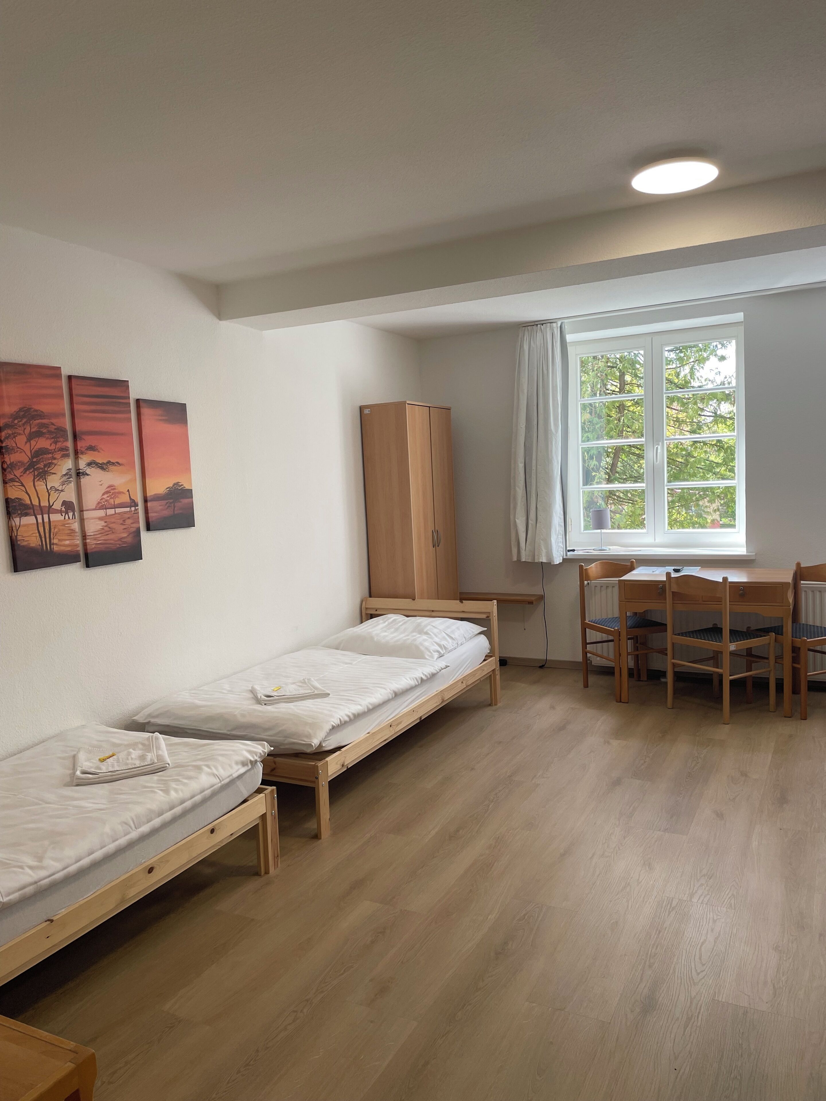 Una habitación con dos camas individuales, un armario de madera, una mesa con sillas y una ventana con vistas a árboles.