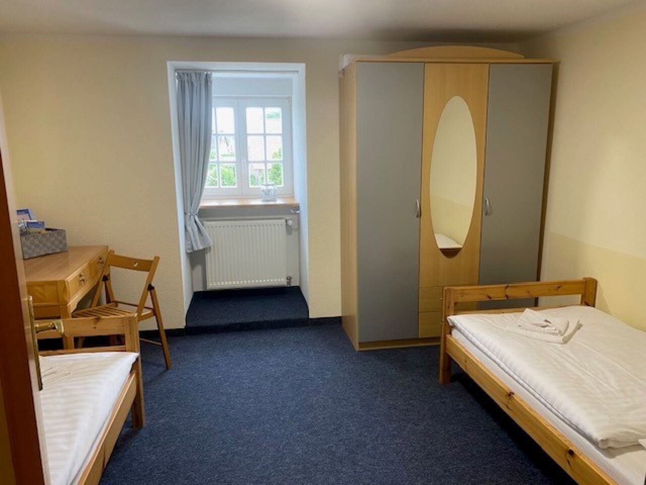 Ein Zimmer mit zwei Einzelbetten, einem Holztisch mit Stuhl, einem Kleiderschrank und einem Fenster mit Vorhängen.