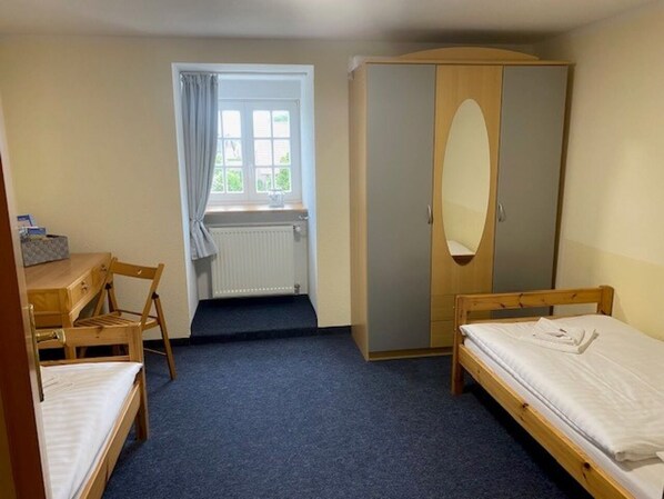 Una habitación con dos camas individuales, un escritorio de madera con una silla, un armario y una ventana con cortinas.