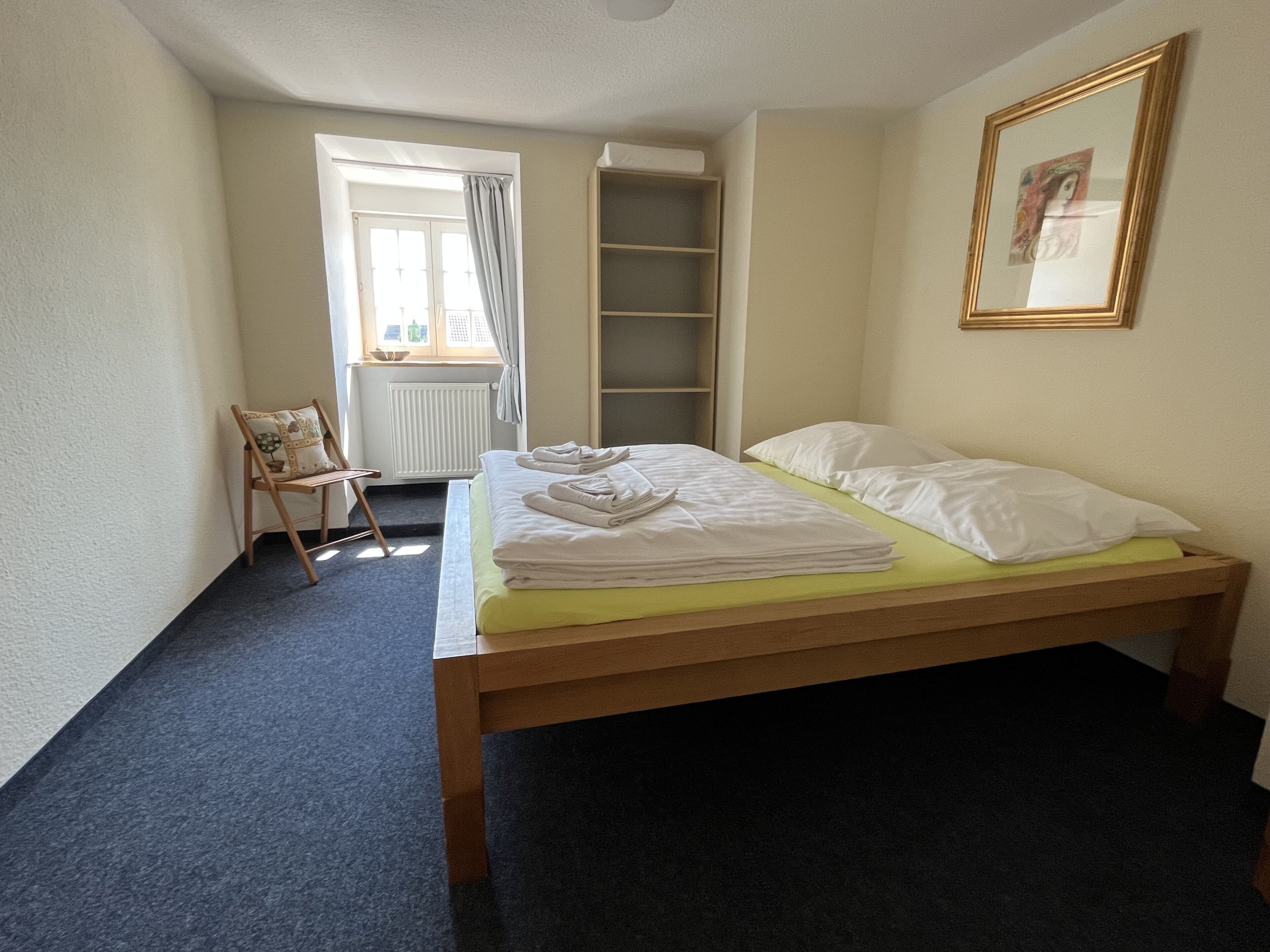 A bedroom with a bed, a chair, a window, and a shelf.